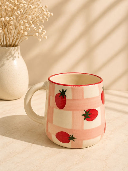 Mug with strawberry pattern on a checkered design, placed on a light surface with a vase of dried flowers in the background.